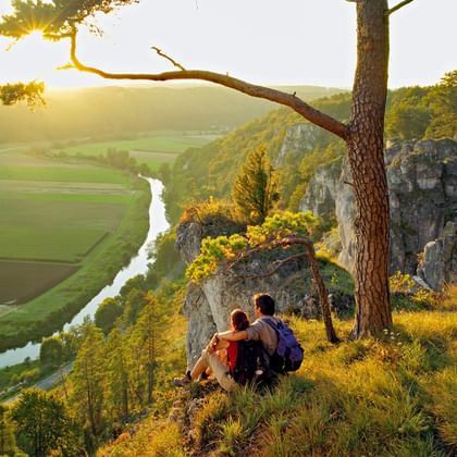 Twee wandelaars zitten op rotsachtige klif met uitzicht over Altmühltal-vallei met kronkelende rivier, groene velden en kalksteenformaties tijdens gouden uur.