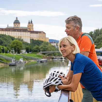 Twee fietsers met helmen genieten van het uitzicht op abdij Melk vanaf de Donau. Het barokke klooster ligt op een heuvel boven de rivier.