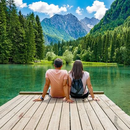 Twee personen zitten op een houten steiger met uitzicht op een turquoise bergmeer omgeven door dicht bos en rotsachtige toppen.