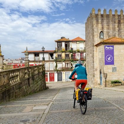 Fietser met gele fietstassen rijdt over kasseien in Porto's historische centrum, langs middeleeuwse stenen toren en kleurrijke huizen.
