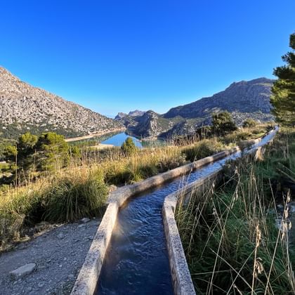 Stenen waterkanaal naar Gorg Blau reservoir in Mallorca's Tramuntana gebergte, omgeven door mediterrane vegetatie en rotsachtige toppen.