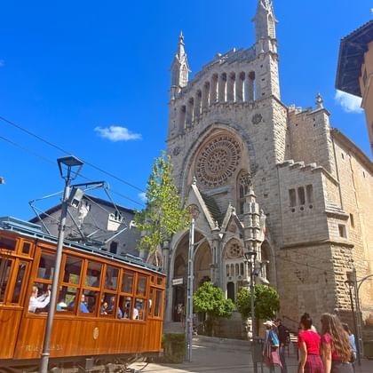Gotische Soller Kathedraal met tweelingtorens en roosvenster op Mallorca. Historische houten tram rijdt voorbij met toeristen onder blauwe lucht.