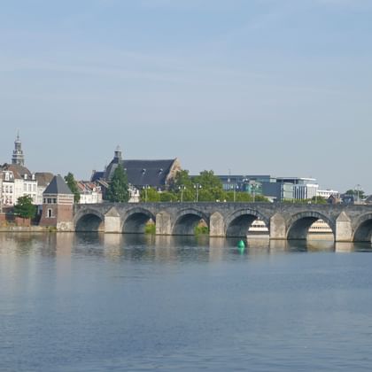 Meerbogige stenen brug over een kalme rivier in Maastricht met historische gebouwen en kerktorens langs de waterkant.