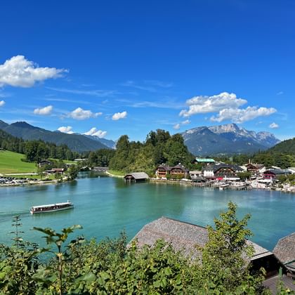 Panoramisch uitzicht op de Königssee met smaragdgroen water, toeristenboot, traditionele Alpengebouwen langs de oever en besneeuwde bergen.