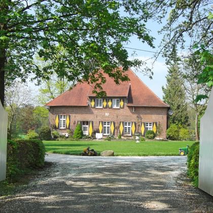 Rueschhaus, een traditioneel bakstenen gebouw met rood pannendak en gele luiken, gezien door witte houten poorten met oprijlaan.