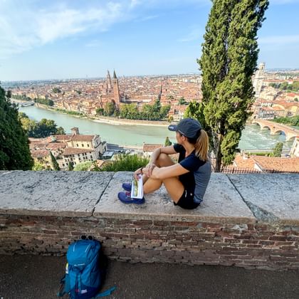 Wandelaar met rugzak zittend op stenen muur met uitzicht op Verona's oude stad, de Adige en historische brug, omlijst door cipressen.