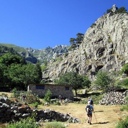 Wandelaar met rugzak op onverhard pad bij Bergerie de Rivisecco op Corsica, met stenen gebouwen en dramatische rotswand onder blauwe lucht.