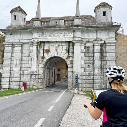 Fietser met witte helm bekijkt een barokke stenen stadspoort in Udine met twee ronde torens en Latijns opschrift boven de boog.