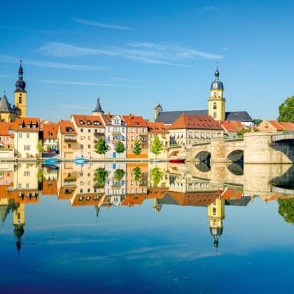 Historische stenen brug over de Main in Kitzingen met kleurrijke gebouwen en kerktorens weerspiegeld in kalm water onder blauwe lucht.