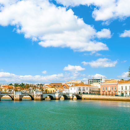 Stenen brug met meerdere bogen over een rivier in Tavira, Algarve. Kleurrijke historische gebouwen langs de waterkant onder een blauwe lucht.