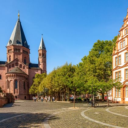 Dom van Mainz met rode zandstenen torens naast een geplaveide plein. Een oranje barok gebouw staat rechts, groene bomen omzomen het plein.