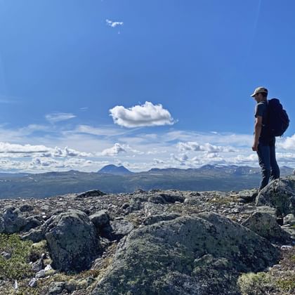 Wandelaar met rugzak staat op rotsachtig terrein in Dovrefjell, Noorwegen, uitkijkend over bergketens onder blauwe hemel met witte wolken.