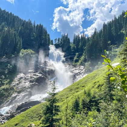 Bovenste Krimml-watervallen die over rotsachtige hellingen vallen, omgeven door groene weiden en dicht bos onder een blauwe lucht.