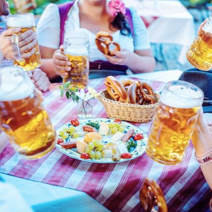 Groep vrienden in traditionele kleding proosten met bierpullen aan een geruite tafel met pretzels en snacks in een biergarten.