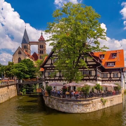 Vakwerkhuis op kanaaleiland in Esslingen met Stadtkirche-torens op achtergrond. Caféterras onder grote boom, blauwe lucht met wolken.