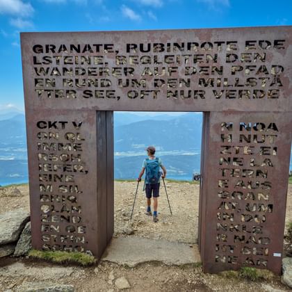 Wandelaar met trekkingstokken loopt door metalen Granattor-poort met uitgesneden tekst op bergtop. Bergketens zichtbaar op achtergrond onder blauwe lucht.