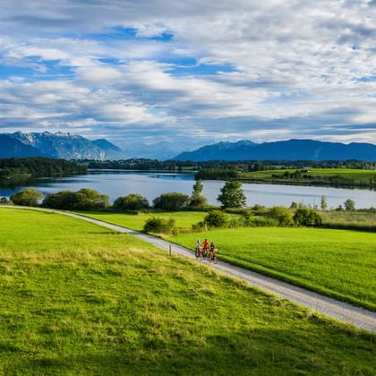 Fietsers op een pad door groene weiden met uitzicht op de Riegsee, met besneeuwde Alpen en bewolkte hemel op de achtergrond.