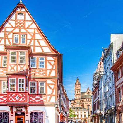 Historische straat in Mainz oude stad met kleurrijke vakwerkhuizen. Rood-wit vakwerkhuis domineert de linkerkant, kathedraal zichtbaar op achtergrond.