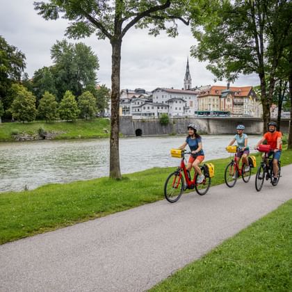 Drie fietsers langs de Isar in Bad Tölz. Historische stad met kerktoren en brug zichtbaar over het water, omlijst door groene bomen.
