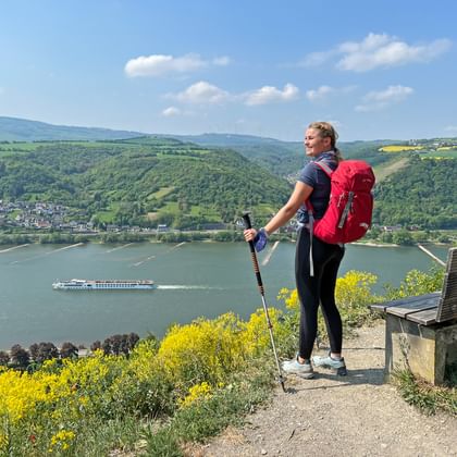 Wandelaar met uitzicht op Rijndal Wandelaarster met rode rugzak en wandelstokken staat op uitkijkpunt met uitzicht op de Rijn met cruiseschip en groene heuvels.