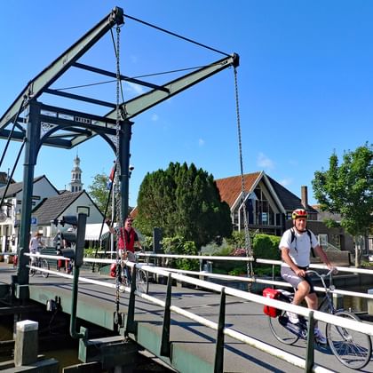 Fietser met fiets staand op een traditionele Nederlandse ophaalbrug over een gracht. Historische gebouwen en kerktoren zichtbaar op de achtergrond onder blauwe lucht.