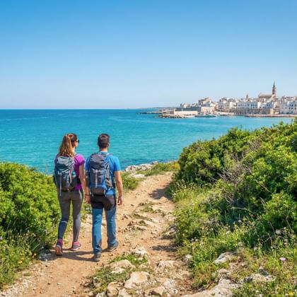 Twee wandelaars met rugzakken op een kustpad in Apulië, met uitzicht op turquoise zee en historische stad met kerktoren.