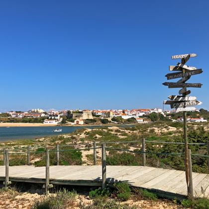 Houten vlonderpad met richtingbord op kustpad met uitzicht op rivier en Vila Nova de Milfontes onder heldere blauwe lucht.
