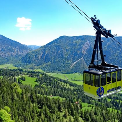 Gele Wendelstein kabelbaan gondel hangt boven beboste bergen en groene valleien in de Beierse Alpen onder blauwe hemel.