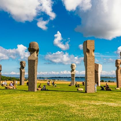 Hoge stenen sculpturen met menselijke gezichten in Dodekalitten park in Kragenæs, Denemarken. Bezoekers ontspannen op groen gazon met kustzicht.