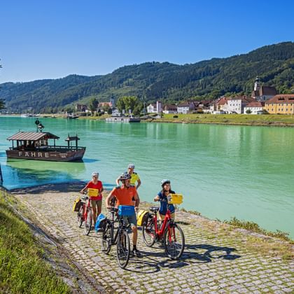 Vier fietsers met fietsen aan rivieroever wachten op veerboot in Engelhartszell. Groene Donau, beboste heuvels en kleurrijke gebouwen zichtbaar.