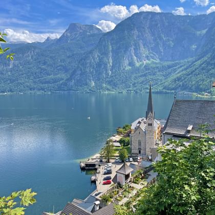 Zicht op Hallstatt kerk met hoge toren aan het meer, omringd door bergen. Blauw meerwater en groene hellingen onder bewolkte hemel.
