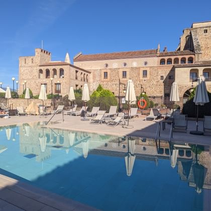 Zwembad bij Parador de Plasencia met historische stenen gebouwen op de achtergrond. Zwembadgebied met ligstoelen en parasols onder blauwe hemel.
