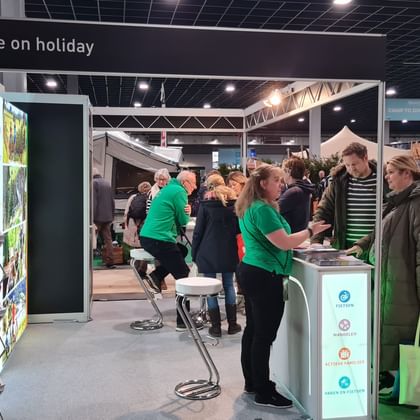 Beurstand van Active on Holiday met personeel in groene shirts die bezoekers helpen. Folderstandaards en informatiebalie zichtbaar.