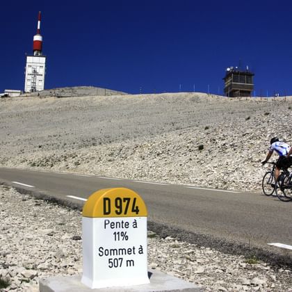 Twee fietsers klimmen Mont Ventoux met afstandspaal 974m naar top bij 11% helling. Witte toren en observatorium zichtbaar op de top.