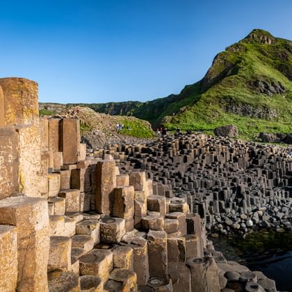 Zeshoekige basaltkolommen bij Giant's Causeway met groene kusthellingen en blauwe lucht. Bezoekers lopen tussen de donkere steenformaties bij het water.