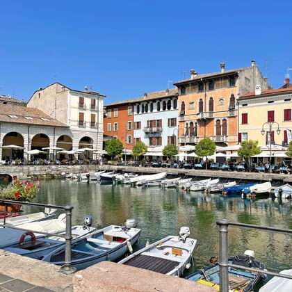 Haven van Desenzano aan het Gardameer met aangemeerde boten, kleurrijke historische gebouwen en witte boogzuilengang onder blauwe lucht.