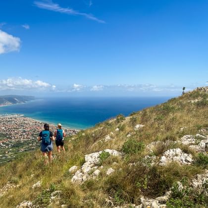 Twee wandelaars op Monte Croce met panoramisch uitzicht op de Ligurische kust en Middellandse Zee. Een topkruis is zichtbaar op de rotsachtige top.