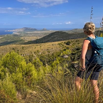 Vrouwelijke wandelaar met blauwe rugzak op heuvel kijkend naar Mallorca's oostkust met turquoise zee en bergachtig landschap.