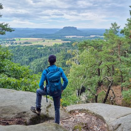 Wandelaar in blauwe jas met rugzak op rotsachtig uitkijkpunt met uitzicht op bebost dal en verre tafelberg in Saksisch Zwitserland.
