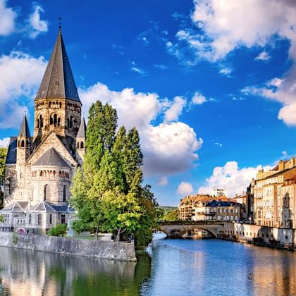 Temple Neuf kerk met hoge toren aan de Moezel in Metz, met historische gebouwen en stenen brug onder blauwe lucht met witte wolken.