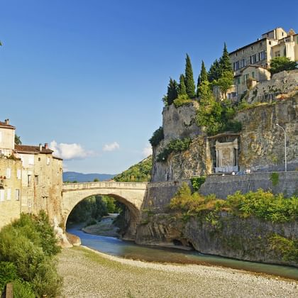 Oude Romeinse stenen brug over rivier in Vaison-la-Romaine met historische gebouwen op heuvel en cipressen onder blauwe hemel.