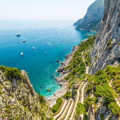 Luchtfoto van Via Krupp, een kronkelend zigzagpad uitgehouwen in steile kliffen op Capri, afdalend naar turquoise Middellandse Zee met boten.