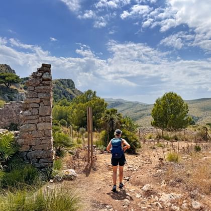 Wandelaar met rugzak verkent de stenen ruïnes van Llevant op Mallorca, omringd door mediterraan landschap met heuvels en blauwe lucht.
