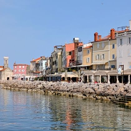 Kleurrijke gebouwen langs de boulevard van Piran in Slovenië met rotsachtige kustlijn en kalme Adriatische Zee onder heldere blauwe lucht.