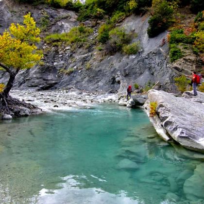 Wandelaars steken turquoise poel over in rotsachtige kloof met herfstbomen. Hidden Valley trek in Albanië toont helder water omringd door grijze rotsen.