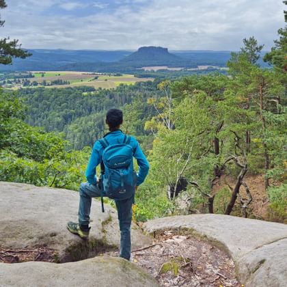 Wandelaar met blauwe rugzak staat op rotsuitstulping en kijkt naar Lilienstein berg over bebost dal vanaf Ochelwände uitkijkpunt.