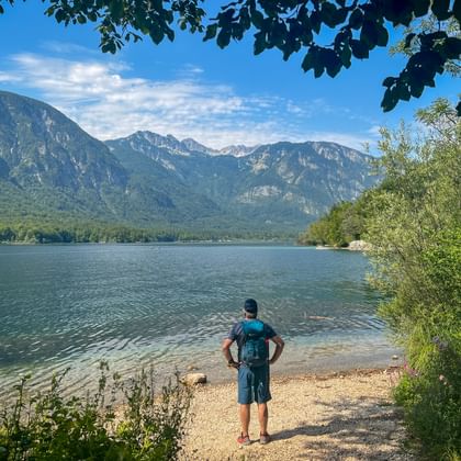Wandelaar met rugzak op kiezelstrand bij het Bohinjmeer, met uitzicht op de bergen van de Julische Alpen onder blauwe lucht met wolken.