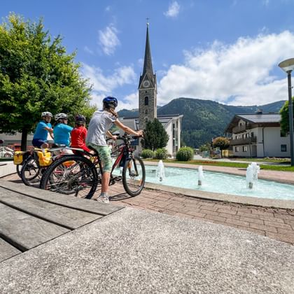 Familie van vier fietsers met fietsen bij fontein op dorpsplein Maishofen, met kerktoren en Alpengebergte op achtergrond.