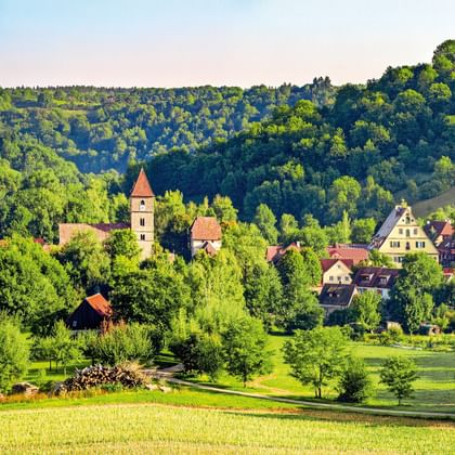 Panoramisch zicht op Rothenburg ob der Tauber met historische torens en gebouwen tussen groene heuvels, bossen en weiden onder blauwe lucht.