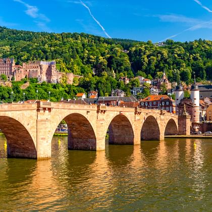 Historische stenen boogbrug over de Neckar in Heidelberg met kasteelruïne op beboste heuvel en kerktoren in de oude stad.